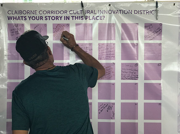 A person writes their thoughts on a sticky note on a board that says "What's your story in this place?"
