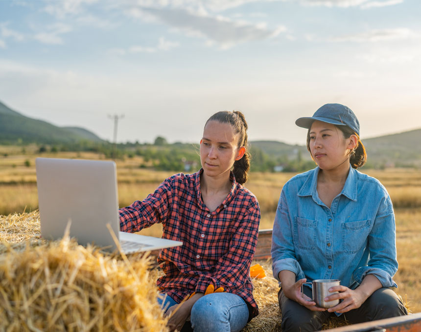 Two women look at a laptop while sitting in the back of a truck on bales of hay.