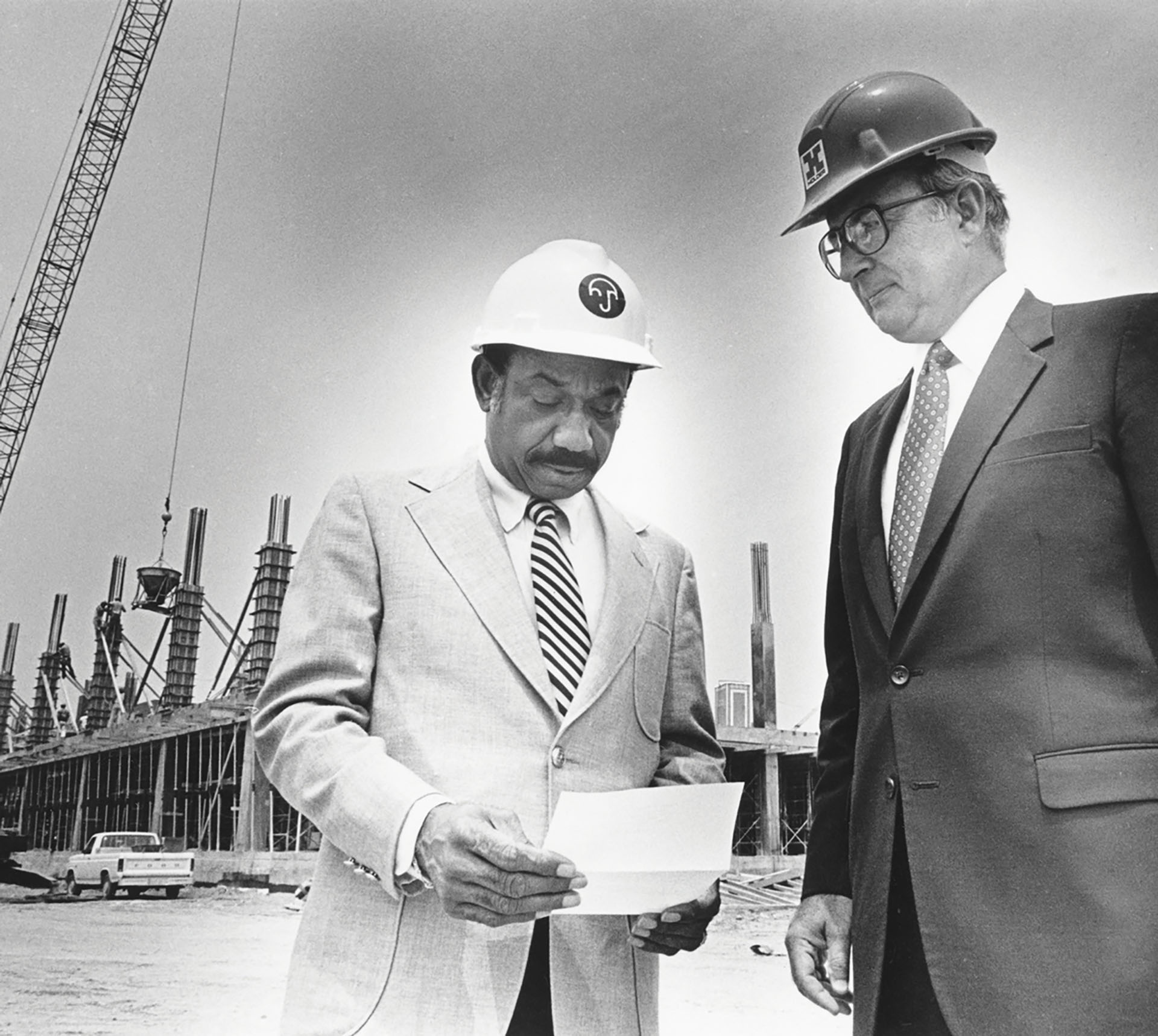 Herman J. Russell and a business associate look over paperwork while wearing hard hats and suits on a construction site