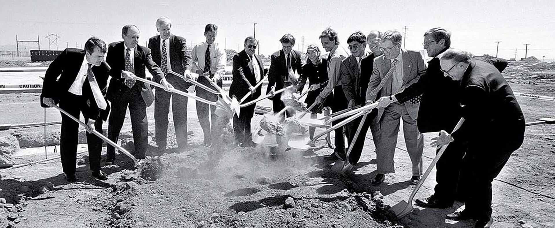 Twelve men and one woman in business clothes shovel dirt ceremonially at a groundbreaking event