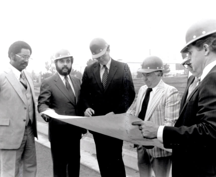 Five men in suits and hardhats look at plans on a construction site
