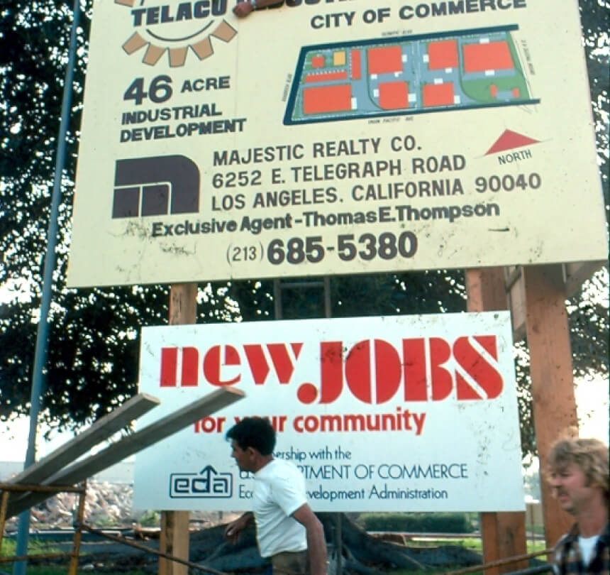 Construction workers walk in front of a sign detailing the construction of the TELACU complex
