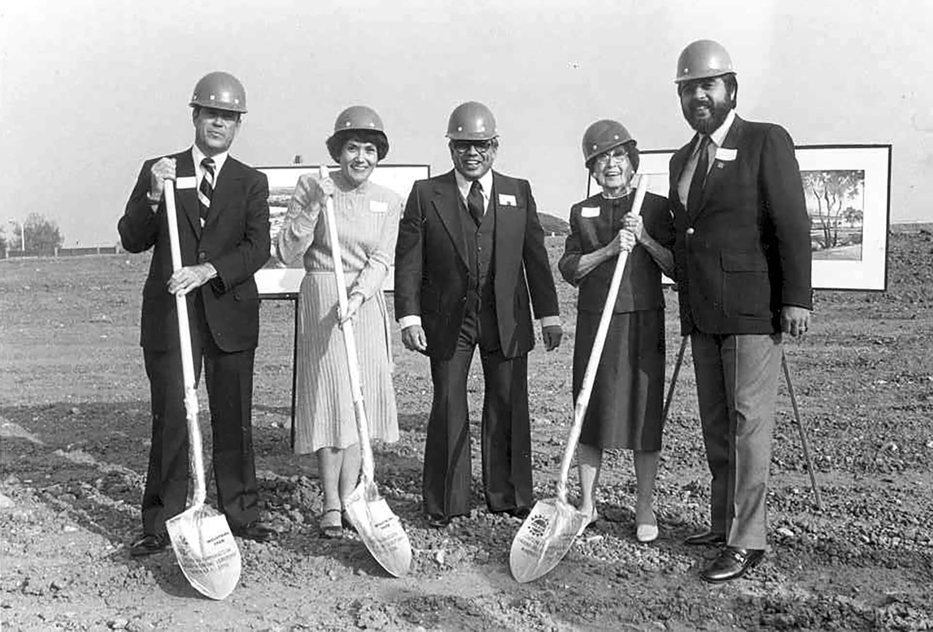 Two women and three men in hard hats and business clothes ceremonially pose with shovels at a groundbreaking event
